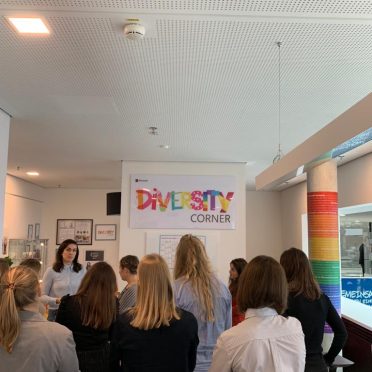 eine Frau erzählt vor einer kleinen Gruppe junger Frauen. Im Hintergrund hängt ein Banner mit der bunten Aufschrift "Diversity Corner"