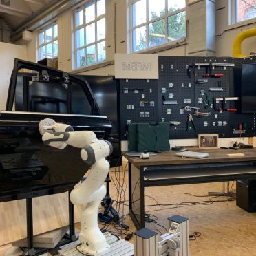 Im Vordergrund befindet sich eine Roboterarm vor einer Autotür - im Hintergrund eine kleine Wand mit verschiedenen Werkzeugen.