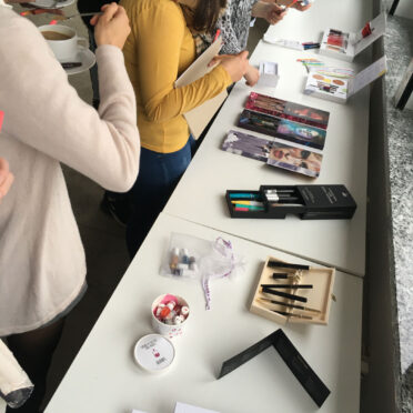 Junge Frauen betrachten diverse Schmink-Utensilien, die auf einem langen Teisch liegen.