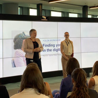 Zwei Frauen halten einen Vortrag vor eine Gruppe - hinter ihnen ein riesiger Bildschirm mit einer Präsentation.