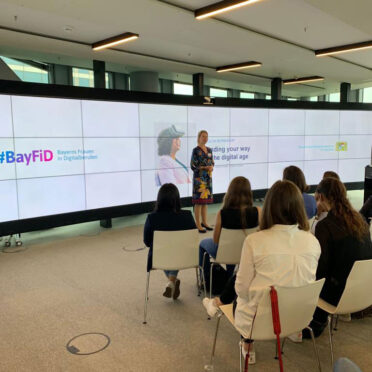 Eine Frau hält einen Vortrag vor eine Gruppe - im Hintergrund eine riesiger Bildschirm mit einer Präsentation.