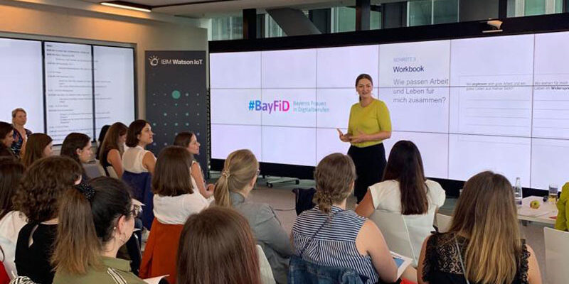 Life Design Workshop in Kooperation mit Yoloa GmbH – Vortrag Judith Gerlach hält einen Vortrag vor eine Gruppe Frauen. Im Hintergrund eine Präsentation, die das BayFiD Logo zeigt.