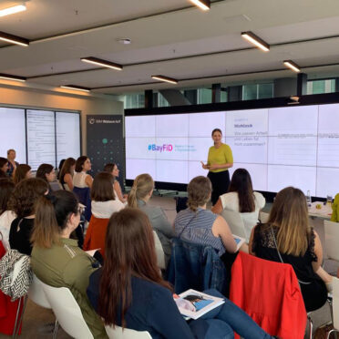 Judith Gerlach hält einen Vortrag vor eine Gruppe Frauen. Im Hintergrund eine Präsentation, die das BayFiD Logo zeigt.