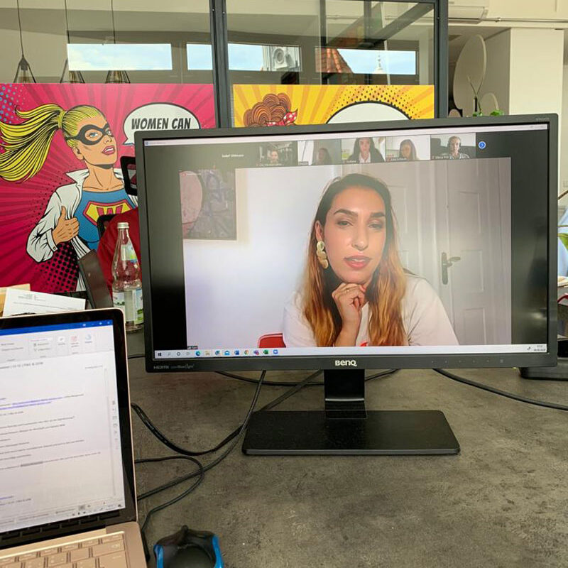 Ein Monitor steht auf dem Boden, daneben ein Laptop. Auf dem Monitor ist Tijen Onaran im Gespräch zu sehen.