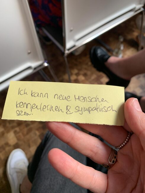 Eine Hand hält einen kleinen Zettel, auf dem steht: "Ich kann neue Menschen kennenlernen und sympathisch sein"